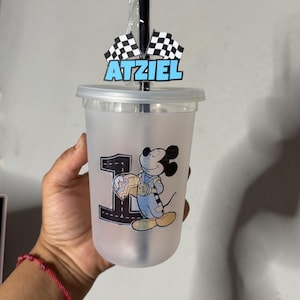 May include: A frosted plastic tumbler with a lid and black straw, featuring a graphic of Mickey Mouse in a racing suit next to a large number one. A topper on the straw displays two checkered racing flags and the name Atziel in blue letters.