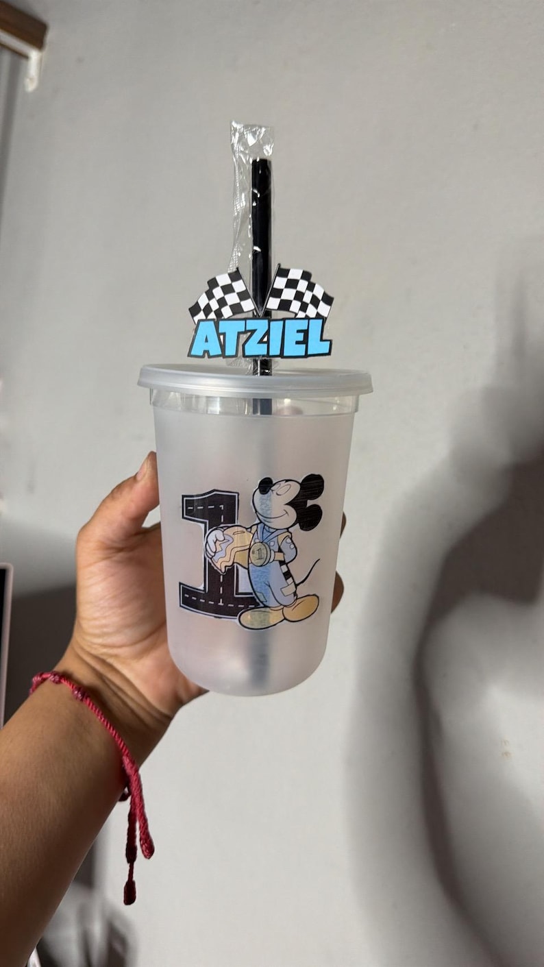 May include: A frosted plastic tumbler with a lid and black straw, featuring a graphic of Mickey Mouse in a racing suit next to a large number one. A topper on the straw displays two checkered racing flags and the name Atziel in blue letters.