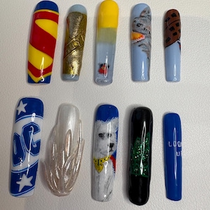 Pode incluir: Um conjunto de dez unhas postiças longas com vários designs. Os designs incluem um padrão amarelo e vermelho, glitter dourado, um gato de desenho animado, um cão e o texto "LOOK UP". Outras unhas apresentam o logotipo da DC e um design perolado.
