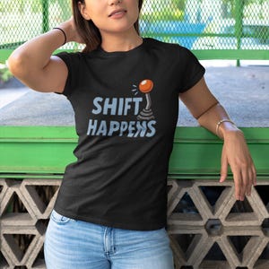Könnte beinhalten: Schwarzes T-Shirt mit dem Schriftzug "SHIFT HAPPENS" in Hellblau und einer Cartoon-Schalthebel-Grafik. Das Model trägt eine hellblaue Jeans. Der Hintergrund ist eine grüne und graue Wand.