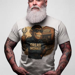 Può includere: T-shirt bianca con un personaggio di scimmia dei cartoni animati con occhiali da sole che tiene una chiave inglese. La scimmia indossa una divisa da meccanico con la scritta "Grease Monkey". Lo sfondo mostra un garage con la scritta "Earhead Goodz".