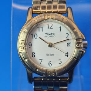 Op de afbeelding: Goudkleurig Timex Indiglo polshorloge met een witte wijzerplaat. Het horloge heeft zwarte cijfers, wijzers en een datumweergave op 3 uur. De horlogeband en kast zijn goudkleurig. Waterbestendig tot 30 meter.