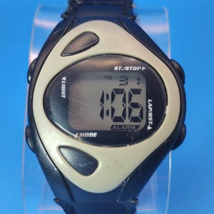 Puede incluir: Un reloj deportivo digital negro y beige con una correa de cuero negro. La esfera muestra la hora, el día y la configuración de la alarma. Funciones: luz, modo, inicio/parada y vuelta/reinicio.