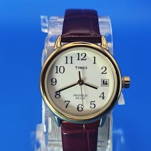 Könnte beinhalten: Eine goldfarbene Timex-Uhr mit weißem Zifferblatt, schwarzen Ziffern und Datumsfenster. Das Armband ist aus weinrotem Leder. Auf dem Zifferblatt steht "Timex" und "Indiglo WR 30M".