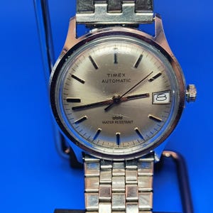 May include: A silver-toned Timex Automatic wristwatch with a silver dial and a date window at the 3 o'clock position. The watch has a stainless steel band and is water resistant. The watch face displays the brand name and the word "Automatic".