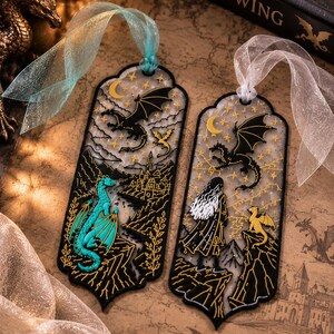 May include: Two rectangular bookmarks with scalloped edges and sheer ribbons. The left bookmark features a turquoise dragon, a black dragon, and a castle. The right bookmark shows a figure with long hair, a black dragon, and a yellow dragon.