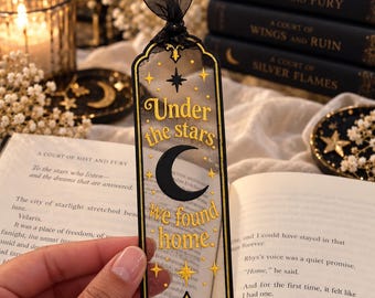 May include: A rectangular, translucent bookmark with a black border and a black ribbon at the top. It features a black crescent moon and gold text that reads, Under the stars, we found home, surrounded by gold star icons and decorative flourishes.