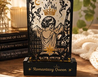 May include: A black rectangular decorative stand featuring a cutout design of a person reading, a crown, dragons, mountains, and the text XIII and The Reader. The base is black with the gold text Romantasy Queen and star accents on each side.