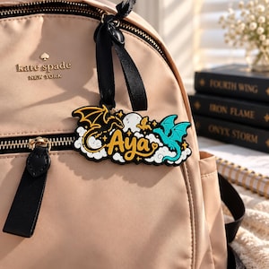 May include: A beige backpack with a black zipper and a black logo tag reading kate spade NEW YORK. Attached to the zipper is a black rubber keychain featuring the name Aya in yellow script, flanked by a black dragon, a teal dragon, and white clouds.