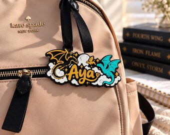 May include: A beige backpack with a black zipper and a black logo tag reading kate spade NEW YORK. Attached to the zipper is a black rubber keychain featuring the name Aya in yellow script, flanked by a black dragon, a teal dragon, and white clouds.