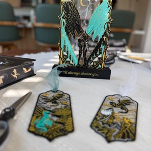 May include: A decorative acrylic stand featuring two dragon heads, a central figure, and the text "I'll always choose you." Two matching bookmarks with dragon illustrations and ribbon tassels are positioned in front of the stand on a white surface.