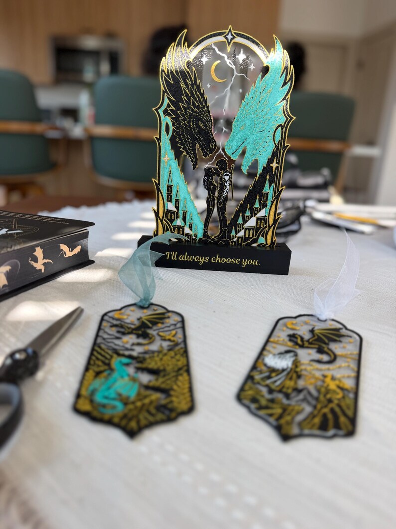 May include: A decorative acrylic stand featuring two dragon heads, a central figure, and the text "I'll always choose you." Two matching bookmarks with dragon illustrations and ribbon tassels are positioned in front of the stand on a white surface.