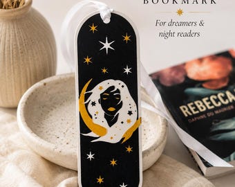 May include: A vertical bookmark with a black background featuring a white silhouette of a face surrounded by a yellow crescent moon and scattered white and yellow stars. A white ribbon is attached to the top. The text reads Stargazer BOOKMARK.