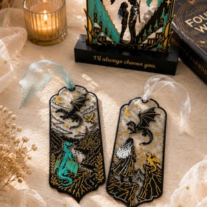 May include: Two rectangular bookmarks with rounded edges and ribbons, featuring black outlines, gold stars, and dragon illustrations. One bookmark shows a teal dragon, the other a person with long hair and a small yellow dragon. Both have intricate mountain scenes.