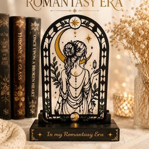 May include: An arched decorative light featuring a black frame with a stained-glass style illustration of a person with an updo hairstyle, a crescent moon, and stars. The base is black with the text In my Romantasy Era printed in gold lettering.