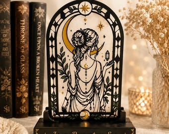 May include: An arched decorative light featuring a black frame with a stained-glass style illustration of a person with an updo hairstyle, a crescent moon, and stars. The base is black with the text In my Romantasy Era printed in gold lettering.