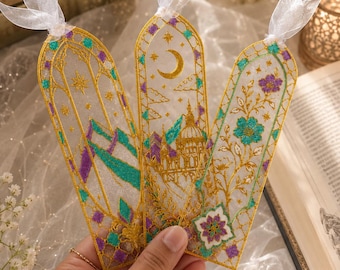 May include: Three translucent bookmarks featuring intricate gold embroidery, teal and purple accents, and white organza ribbon toppers. The designs include arched architectural motifs, a crescent moon, stars, and floral patterns on a sheer base.