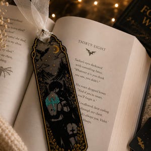 May include: A rectangular bookmark featuring a dark illustration of two figures embracing, accented with a teal dragon tattoo design, a yellow crescent moon, and gold leaf borders. The bookmark is topped with a white sheer ribbon tie.