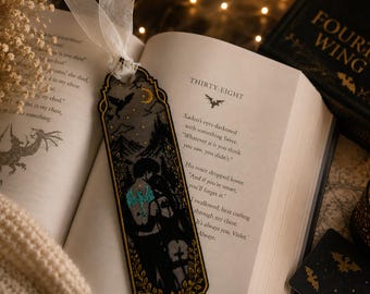 May include: A rectangular bookmark featuring a dark illustration of two figures embracing, accented with a teal dragon tattoo design, a yellow crescent moon, and gold leaf borders. The bookmark is topped with a white sheer ribbon tie.