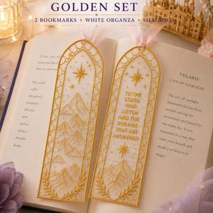 May include: Two rectangular bookmarks with gold embroidered mountain and star designs on white organza fabric. One bookmark features the text "TO THE STARS WHO LISTEN AND THE DREAMS THAT ARE ANSWERED" in gold thread. Both have pink ribbon toppers.