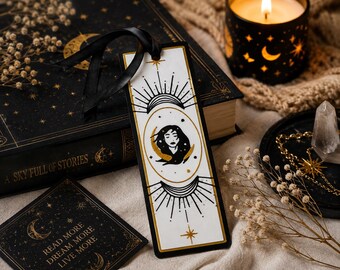 May include: A rectangular white bookmark featuring a black and gold illustration of a person's face surrounded by a crescent moon and sunburst motifs. The bookmark is topped with a black ribbon and displays intricate celestial line art designs.