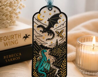 May include: A rectangular bookmark with a scalloped top and bottom, featuring a black laser-cut frame. The design depicts a teal dragon perched on a cliff, a black dragon flying above, a yellow crescent moon, stars, and a castle, finished with a blue ribbon.