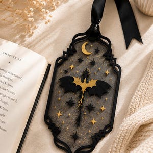 May include: A rectangular black bookmark with an ornate frame, featuring a central gold dragon silhouette surrounded by gold and black stars and a crescent moon. The bookmark is made of a semi-transparent material and includes a black ribbon at the top.