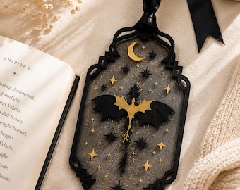 May include: A rectangular black bookmark with an ornate frame, featuring a central gold dragon silhouette surrounded by gold and black stars and a crescent moon. The bookmark is made of a semi-transparent material and includes a black ribbon at the top.