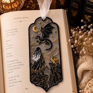 May include: A black and gold metal bookmark featuring a cutout design of a person standing on a cliff looking at a large dragon in the sky and a smaller dragon on a nearby peak, accented with a crescent moon, stars, and a white sheer ribbon topper.