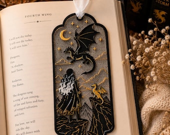 May include: A black and gold metal bookmark featuring a cutout design of a person standing on a cliff looking at a large dragon in the sky and a smaller dragon on a nearby peak, accented with a crescent moon, stars, and a white sheer ribbon topper.