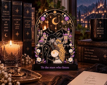May include: An arched decorative light display featuring a black frame with gold and purple accents. The design depicts a couple embracing in front of a castle and mountains under a crescent moon. The base is inscribed with the text To the stars who listen.