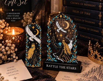 May include: A Throne of Glass gift set featuring a black bookmark with a black ribbon and a matching display stand. Both items feature illustrations of a blonde figure, a white bird, a stag, and a crescent moon. The stand base reads RATTLE THE STARS.