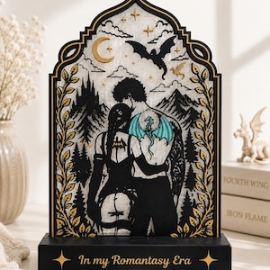 May include: A decorative arched wooden display featuring a black silhouette of two figures embracing, with a teal dragon tattoo on the back of one figure. The background shows mountains, trees, a crescent moon, and stars. The base reads In my Romantasy Era.
