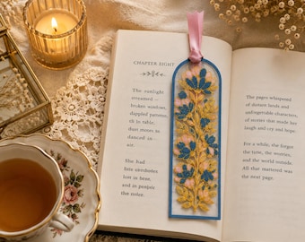 May include: A rectangular bookmark with a rounded top edge, featuring an intricate embroidered floral pattern in shades of blue, pink, and gold on a sheer background. The bookmark is finished with a light pink satin ribbon at the top.