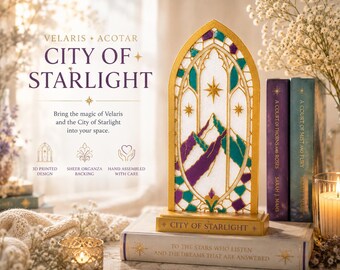 May include: A gold-colored, arched, 3D-printed decorative display piece featuring a mountain range design in purple and teal, with sheer organza backing. The base is inscribed with the text CITY OF STARLIGHT, and it sits atop a book.