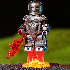 Puede incluir: Figura de Iron Man plateada con casco, armadura de pecho y piernas. La figura se representa con un efecto de llama roja que emana de sus pies y una explosión de energía roja en su mano. El fondo es verde borroso.