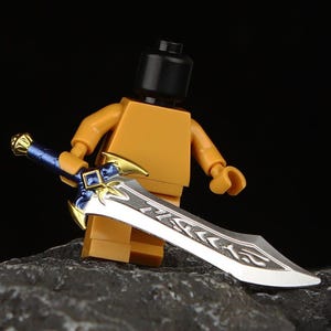 Puede incluir: Una figura de Lego sosteniendo una espada plateada y azul con detalles dorados. La espada tiene un diseño detallado en la hoja y la empuñadura. La figura es naranja y tiene una cabeza negra. El fondo es oscuro.