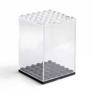 May include: Transparent acrylic display case, shaped like a large building block. The top has clear studs, and the base is dark grey. The cube-shaped case is suitable for showcasing small items, with dimensions in centimetres.