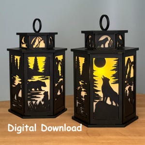 May include: Two black, hexagonal lanterns with cutout forest and animal designs. One lantern features a bear and cubs, the other a howling wolf under a full moon. Each has a handle and is illuminated with a warm glow.