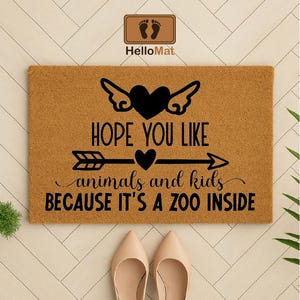 May include: A rectangular doormat with the text "HOPE YOU LIKE animals and kids BECAUSE IT'S A ZOO INSIDE" in black. The design includes a heart with wings, an arrow, and a heart. The mat is brown.