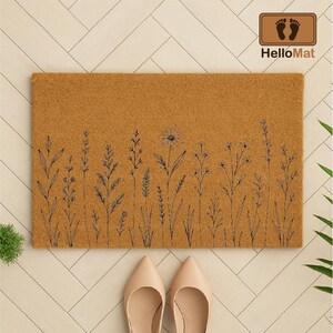 May include: A rectangular doormat with a tan background and a black floral design along the bottom edge. The doormat has the words "HelloMat" printed on a brown rectangular label in the upper right corner.