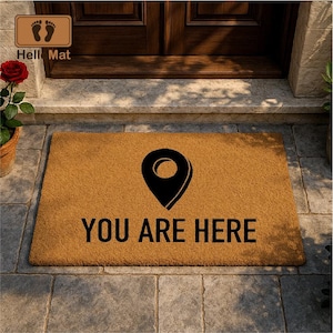May include: A rectangular doormat made of natural fibers, featuring a black location pin graphic and the words "YOU ARE HERE" in bold, black letters. The mat is tan and sits on a stone surface.