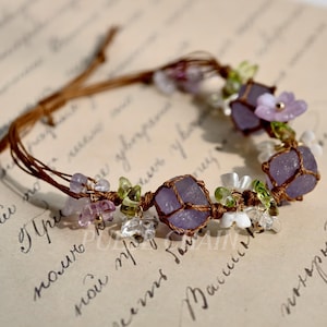 May include: A handmade bracelet featuring lavender-colored stones wrapped in brown cord. The bracelet is adorned with small, clear, green, and purple beads, and tiny flower accents. The bracelet is set against a background of handwritten script.