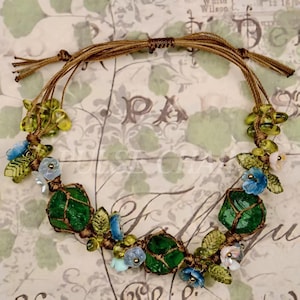 May include: A handmade necklace featuring large, green, irregularly shaped stones wrapped in brown twine. The necklace is adorned with small blue and white flowers, green leaves, and yellow beads. The background is a vintage-style paper with floral and script elements.
