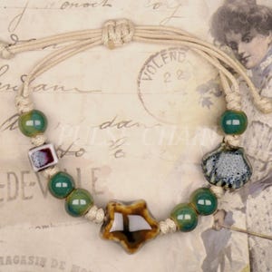 May include: Adjustable bracelet featuring green and brown ceramic beads, a star-shaped charm, and a seashell charm. The bracelet is strung on a beige cord with a sliding knot closure. The bracelet's design incorporates a mix of shapes and colors.