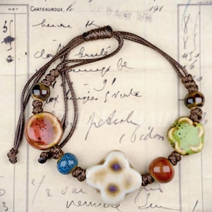 May include: A handmade bracelet featuring a variety of ceramic beads in shades of brown, red, blue, and green. The bracelet includes a four-petal flower-shaped bead and is strung on a brown cord.
