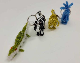 May include: A collection of beaded animal keychains. The keychains include a green and white alligator, a black and white zebra, a gold-colored lion, and a blue starfish. Each keychain has a silver metal ring.