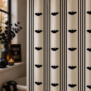 Op de afbeelding: Beige en zwart gestreepte douchegordijn met herhaalde zwarte vleermuis silhouetten. Het gordijn hangt voor een raam. Thema: Halloween.