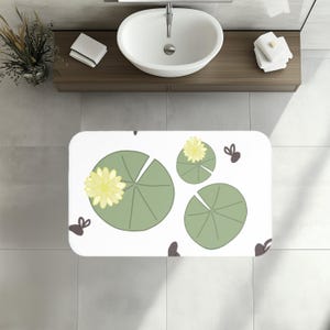 May include: White rectangular bath mat with a lily pad design. The mat features green lily pads with yellow flowers and dark brown accents. The mat is in a bathroom setting.