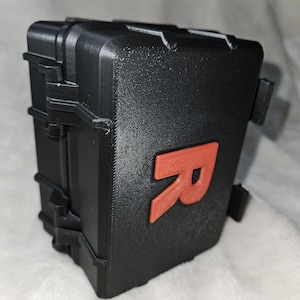 May include: A black, rectangular case with a textured surface and a bright orange letter "R" on the front. The case has a hinged lid and clasps. The overall design suggests a protective container.